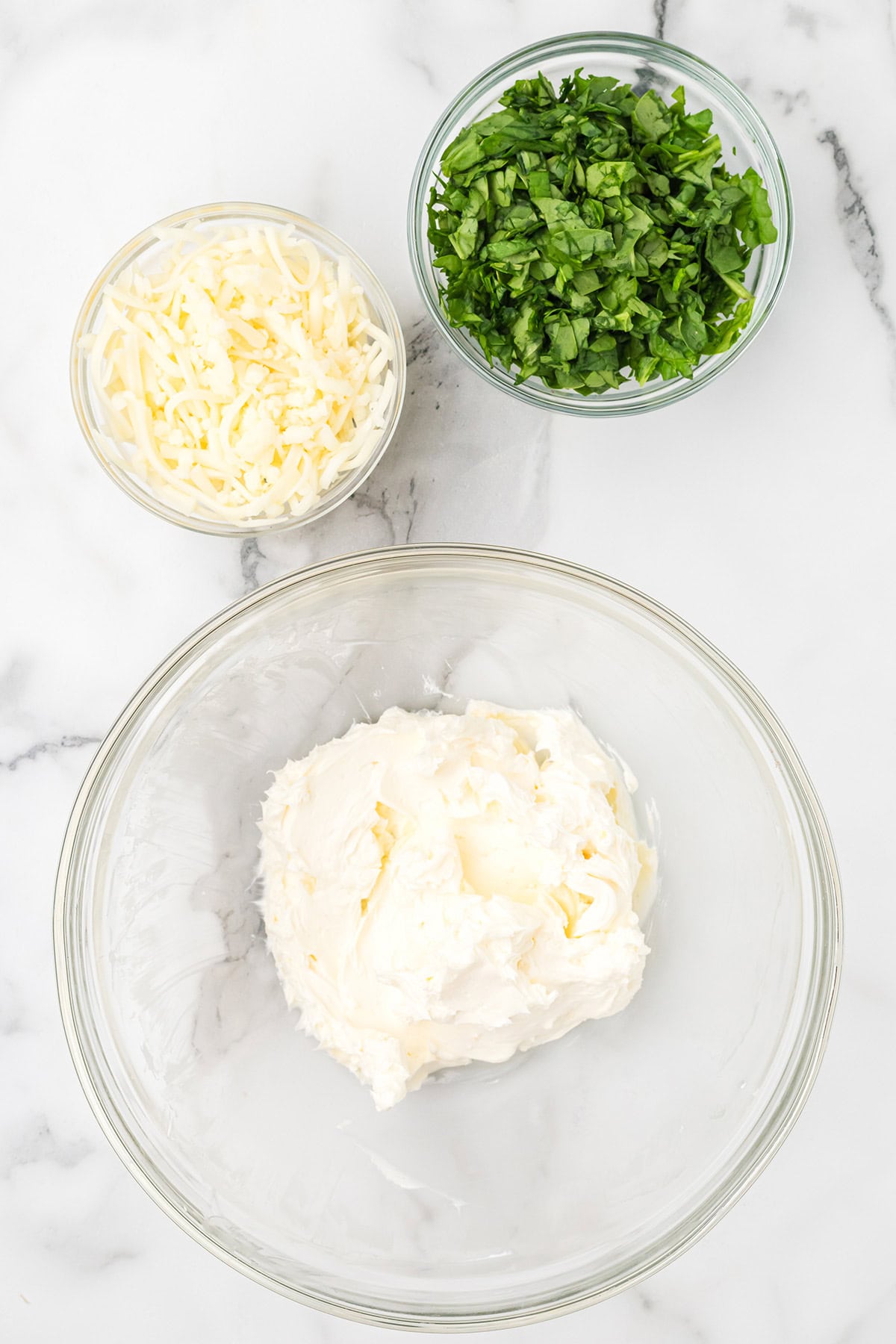 Three bowls with shredded cheese, chopped herbs, and cream cheese on a white marble surface.