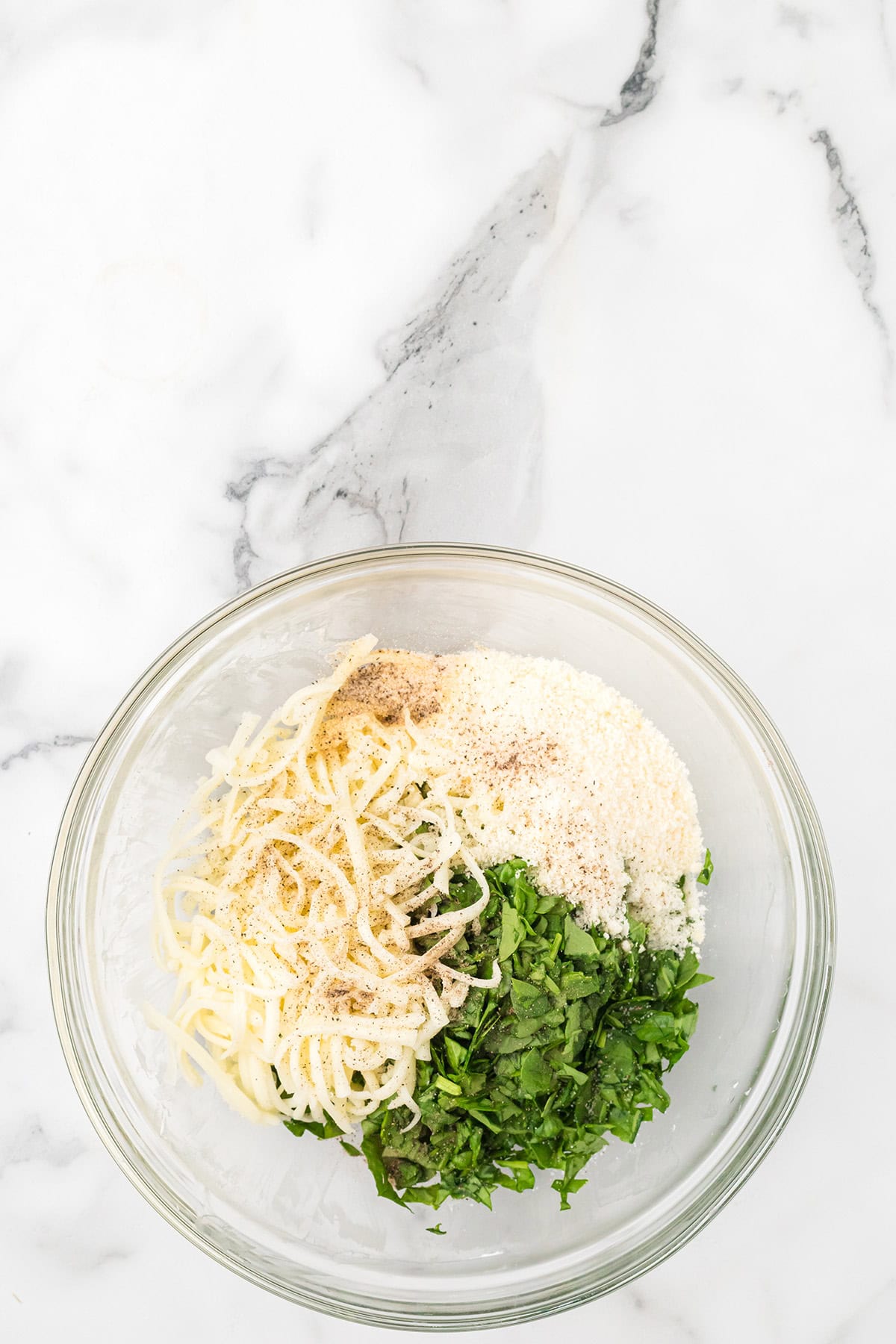 A glass bowl with shredded cheese, chopped herbs, grated parmesan, and ground black pepper on a marble surface.