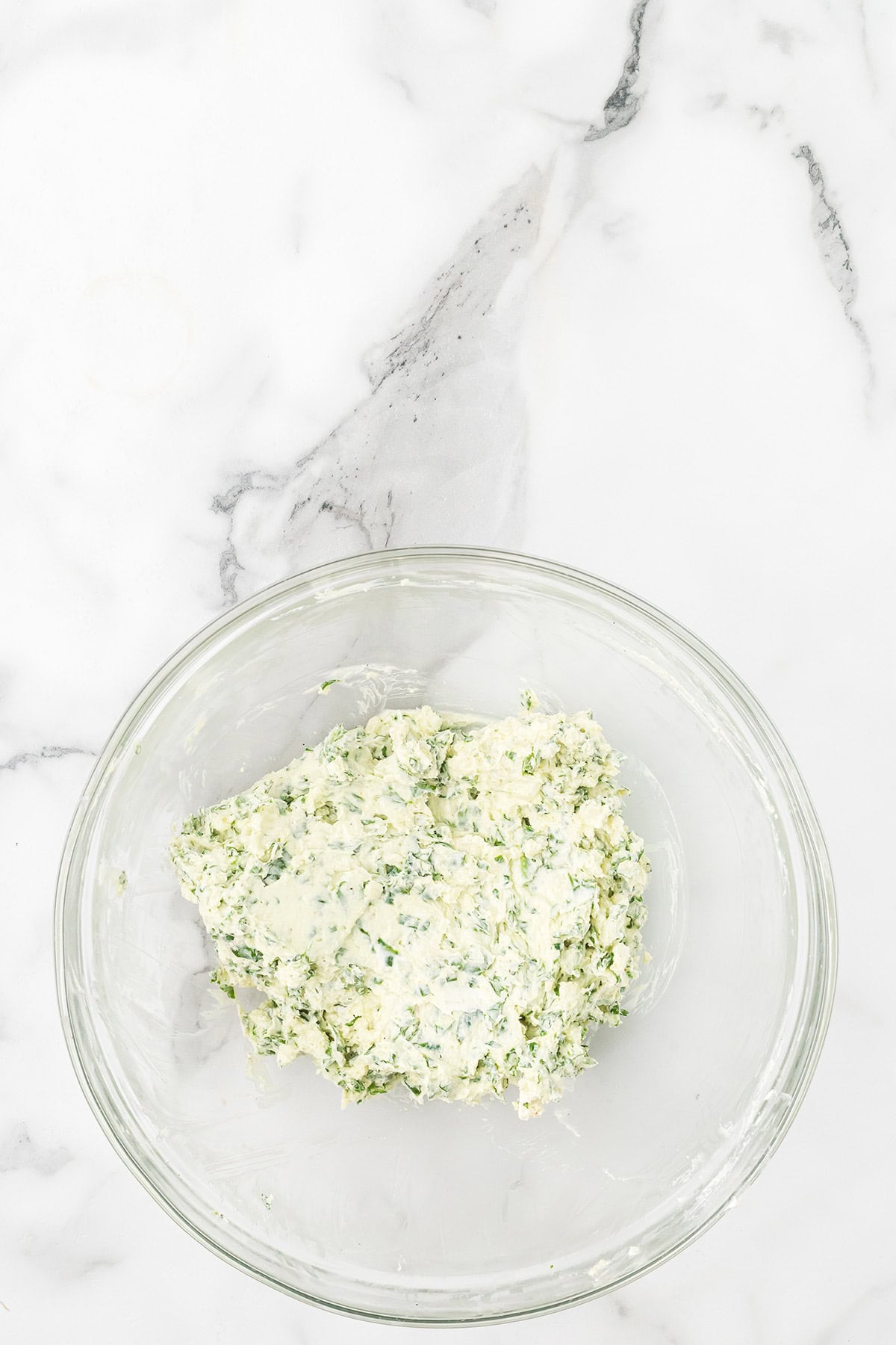 A glass bowl with a mixture of chopped herbs and creamy spread on a white marble surface.