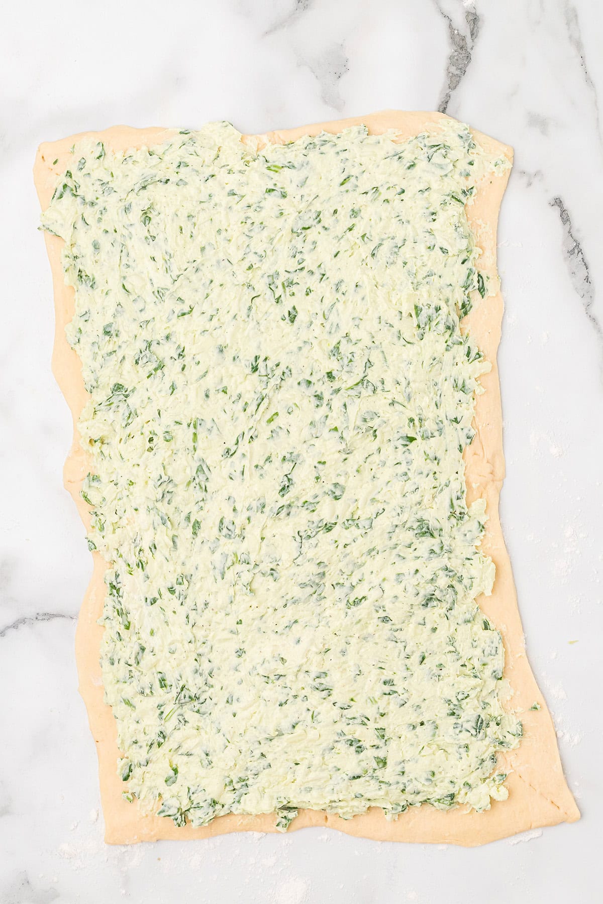 Rectangular dough topped with a cheese and spinach mixture on a marble surface.