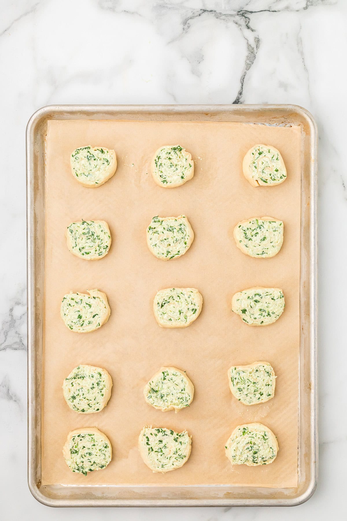 A baking sheet with 15 uncooked spinach pinwheels on parchment paper, on a marble surface.