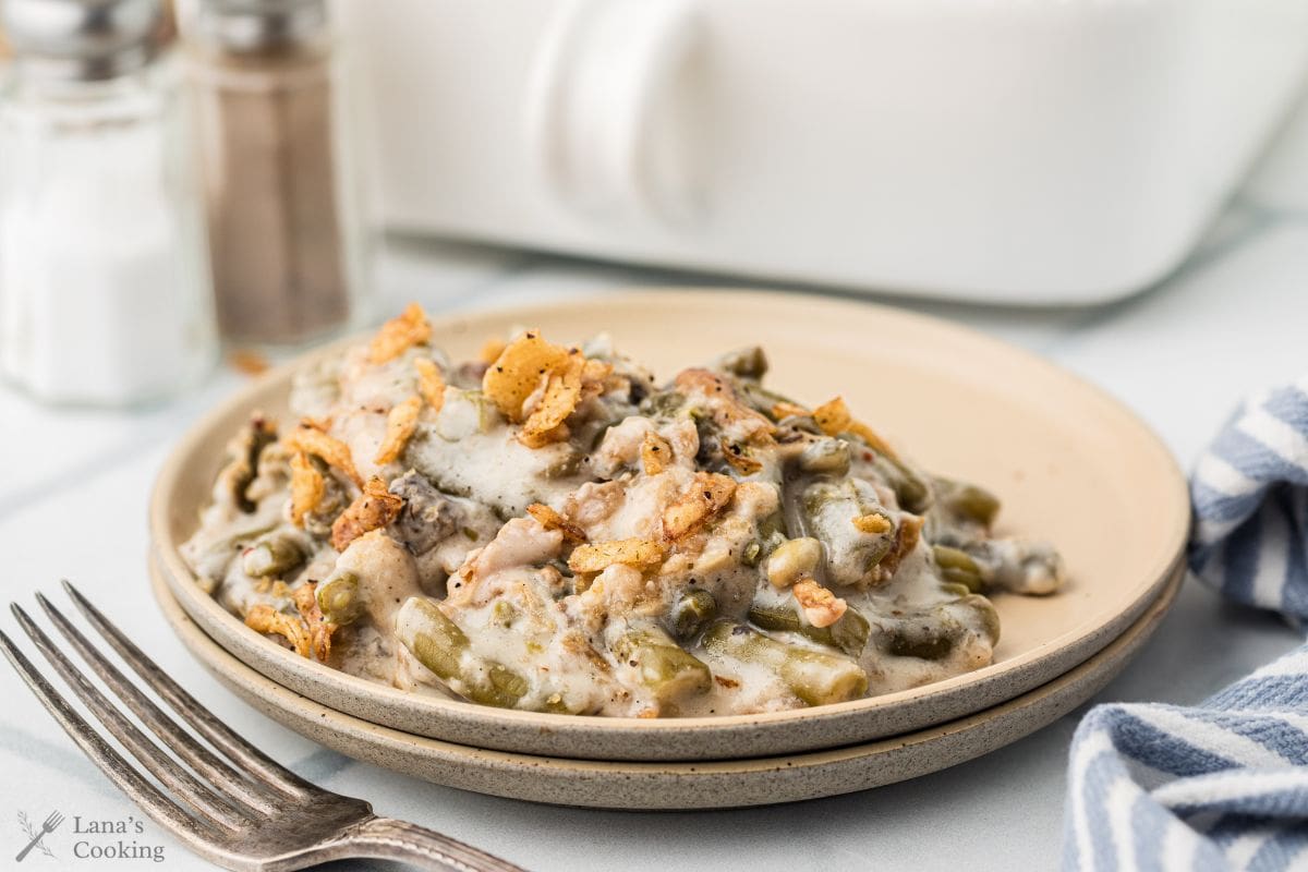 A serving of green bean casserole with crispy onions on a beige plate, next to a fork and napkin.