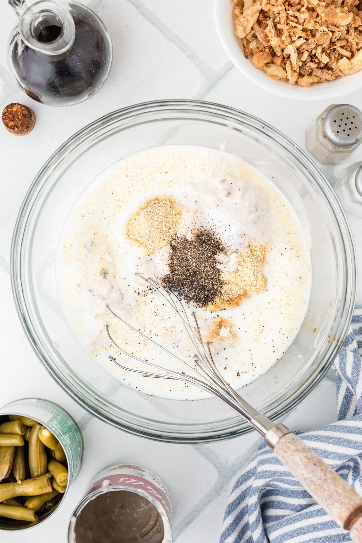 A mixing bowl with milk, spices, and a whisk, surrounded by canned beans and fried onions on a countertop.