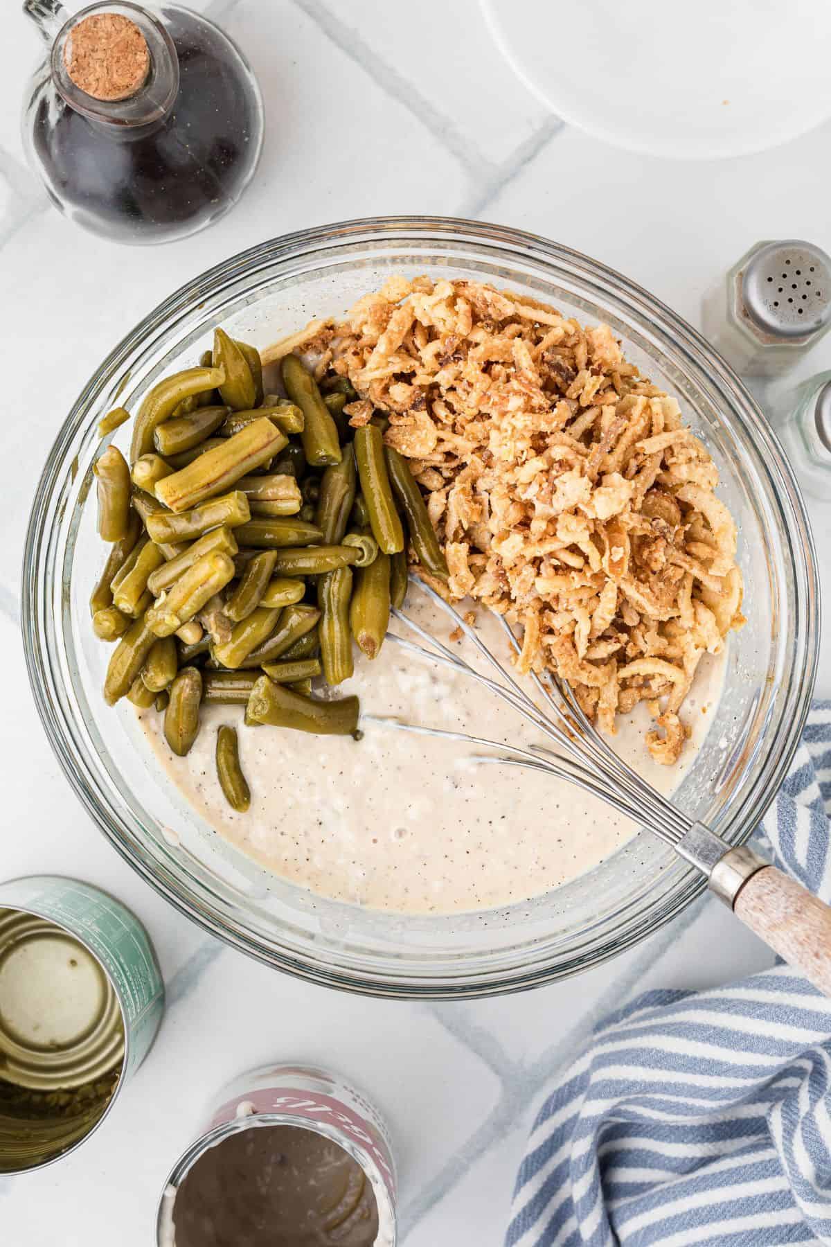 A glass bowl with green beans, crispy onions, and creamy mixture being whisked together on a white surface.