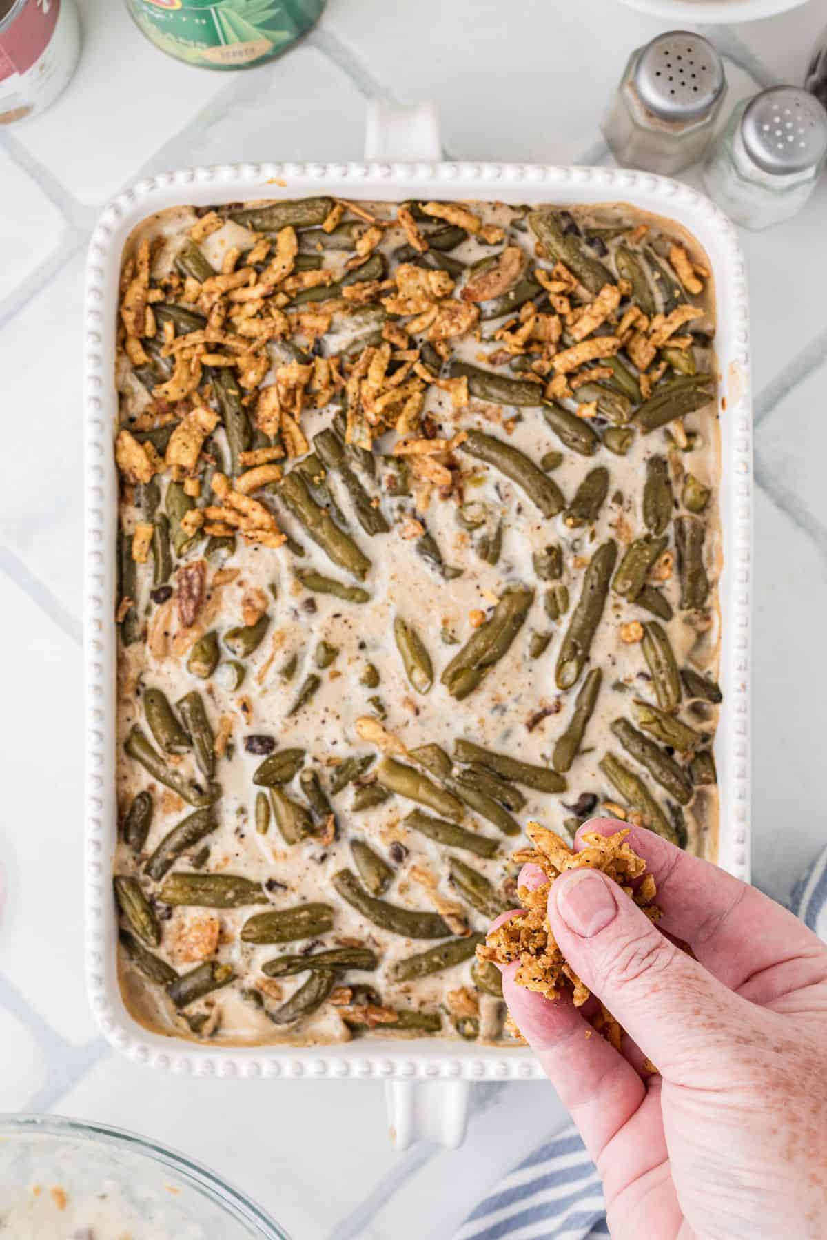 A hand sprinkles crispy onions on a casserole dish filled with green bean casserole.