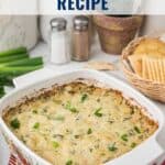 A baked artichoke dip in a white casserole dish, surrounded by crackers, green onions, and seasonings.