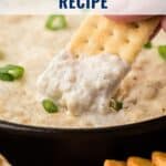 A hand dips a cracker into creamy hot baked crab dip, topped with sliced green onions.