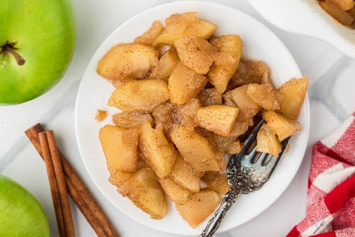 A plate of cooked cinnamon apples with a vintage fork, next to green apples and cinnamon sticks.