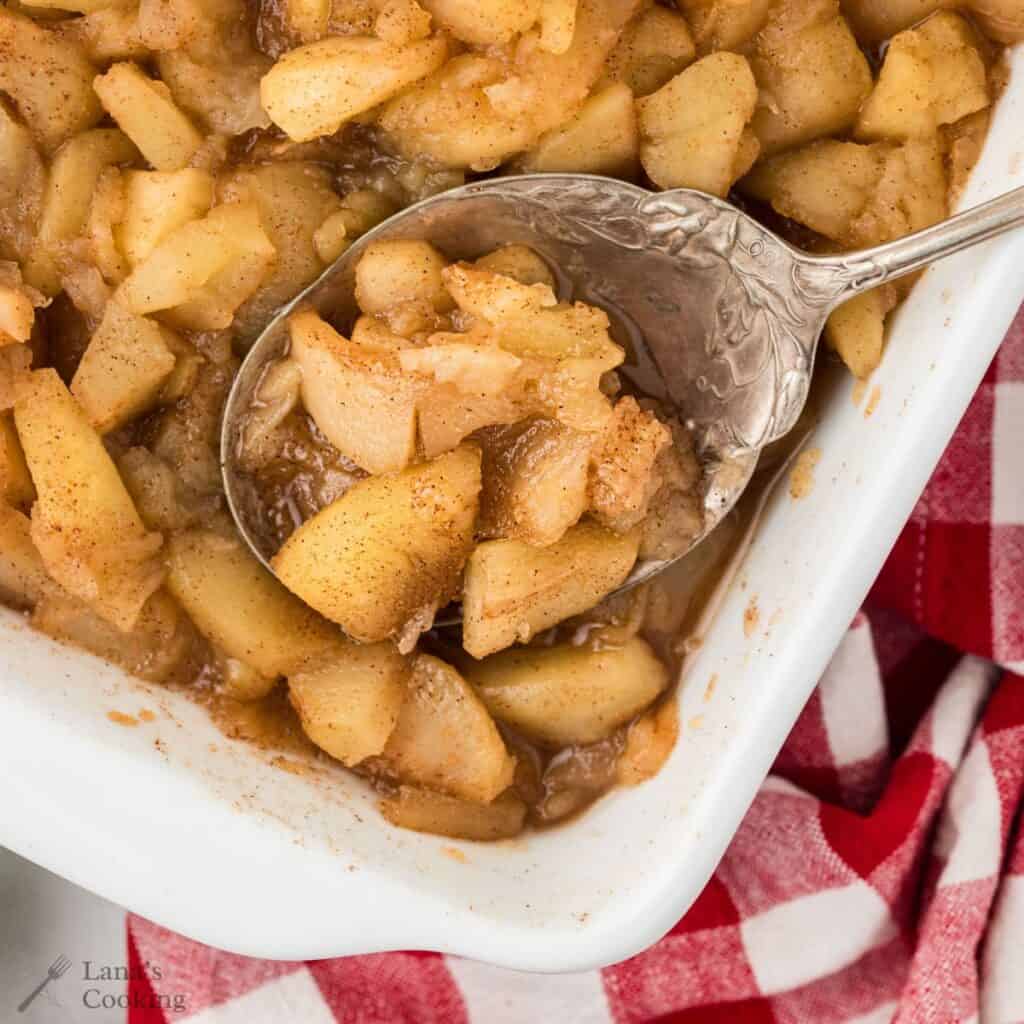 A spoon scoops baked cinnamon apples from a white dish, next to a red and white checkered cloth.