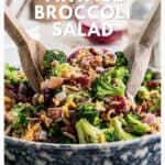A bowl of broccoli salad with wooden servers, topped with bacon, cheese, red onion, and cranberries.