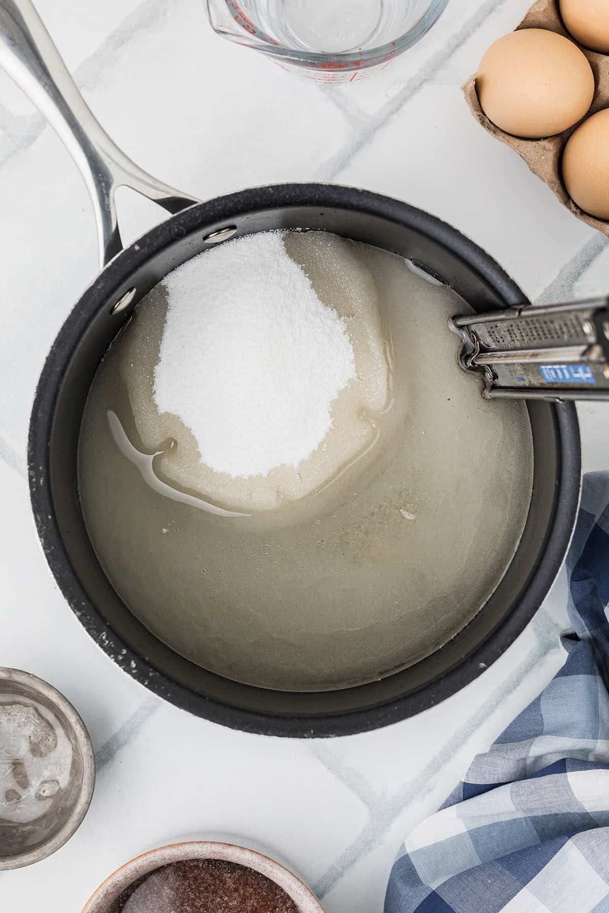A saucepan with sugar, syrup, and water inside, a candy thermometer, and eggs nearby on a marble counter.