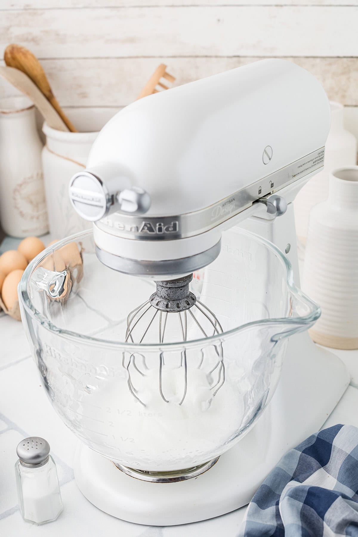 A white stand mixer with a glass bowl sits on a kitchen counter next to eggs, utensils, and a salt shaker.