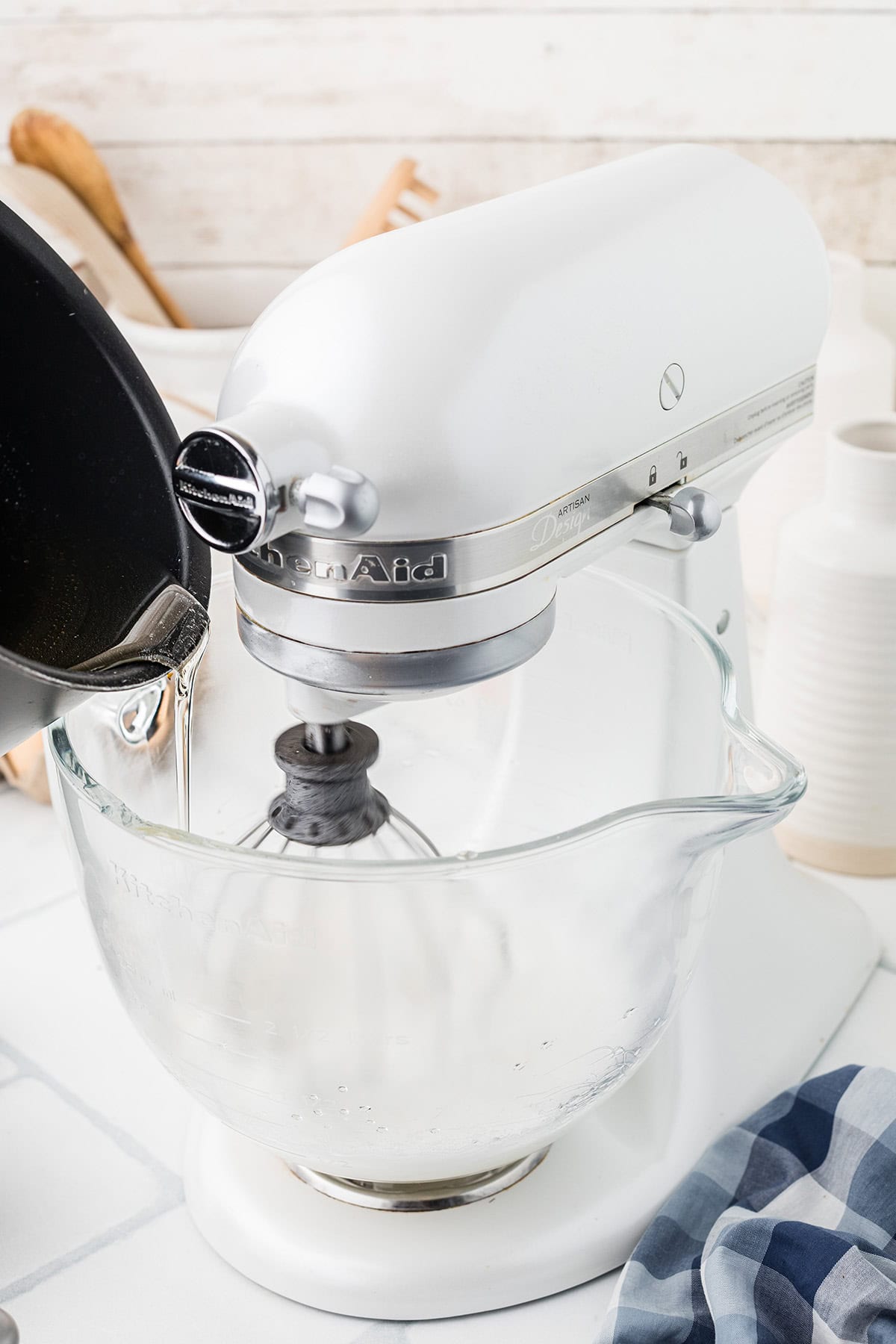 A white stand mixer with a glass bowl as syrup is poured in, with kitchen items in the background.