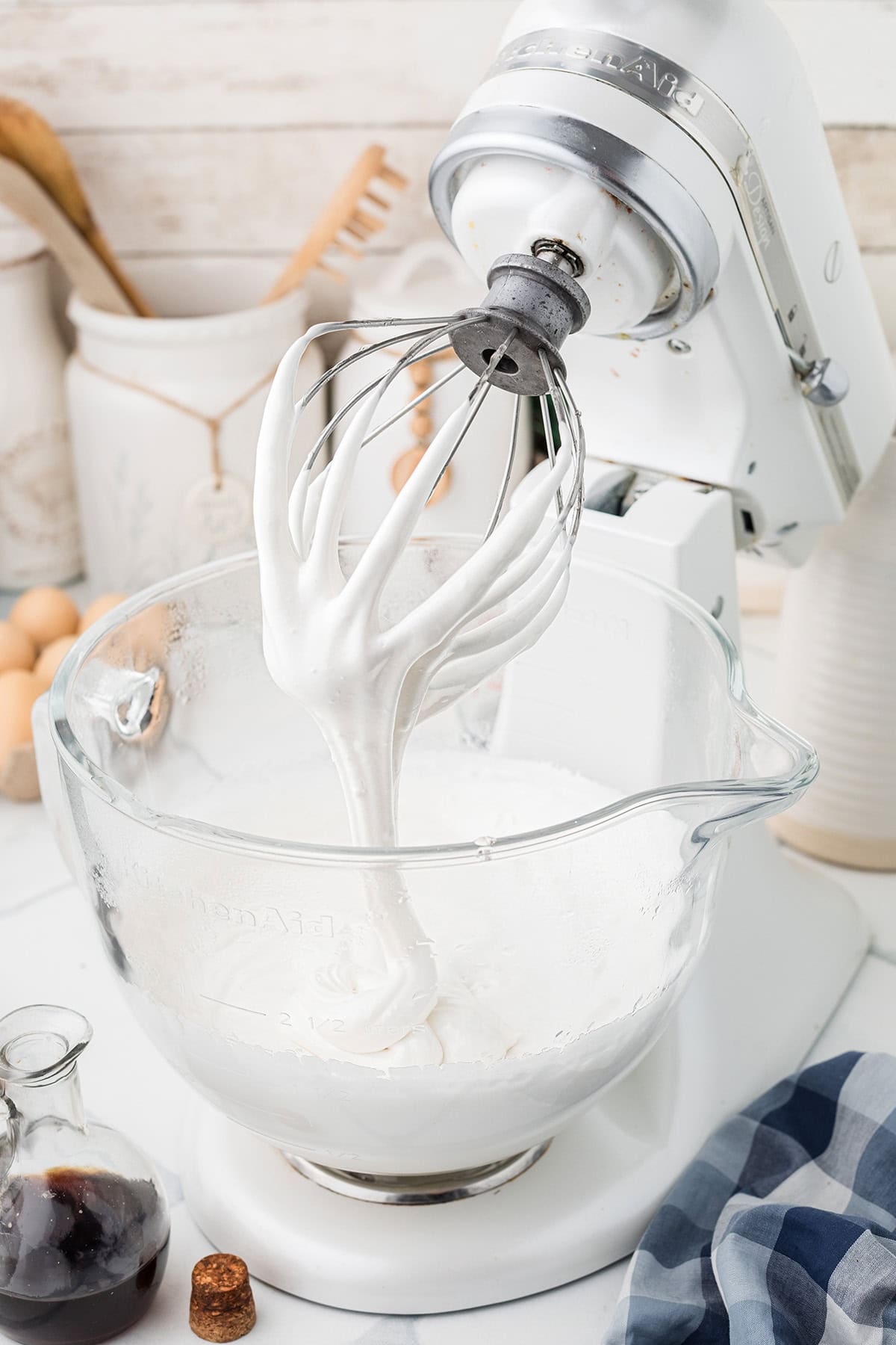 Stand mixer with a whisk attachment covered in fluffy white meringue, next to a glass bowl and ingredients.
