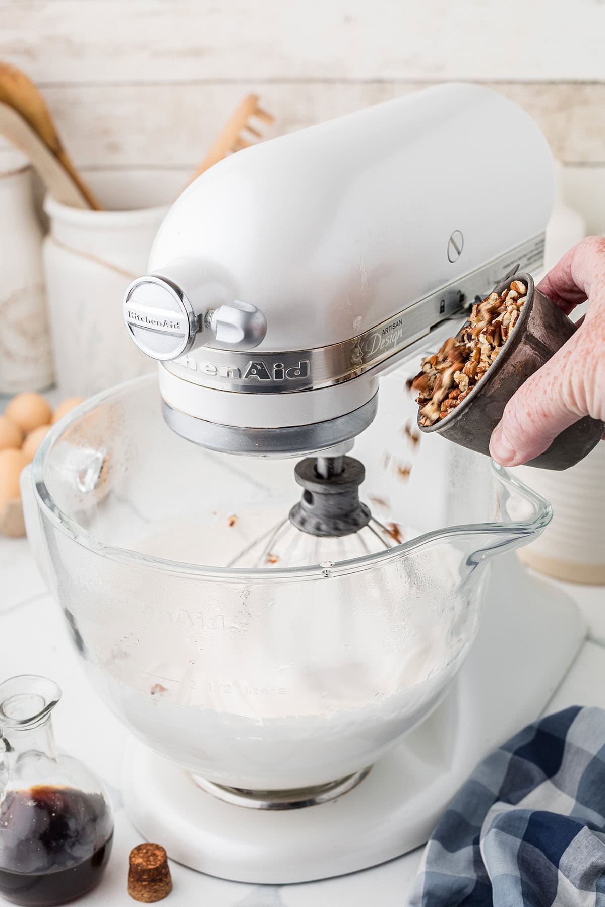 A hand pours chopped nuts into a stand mixer with a glass bowl filled with a creamy mixture.