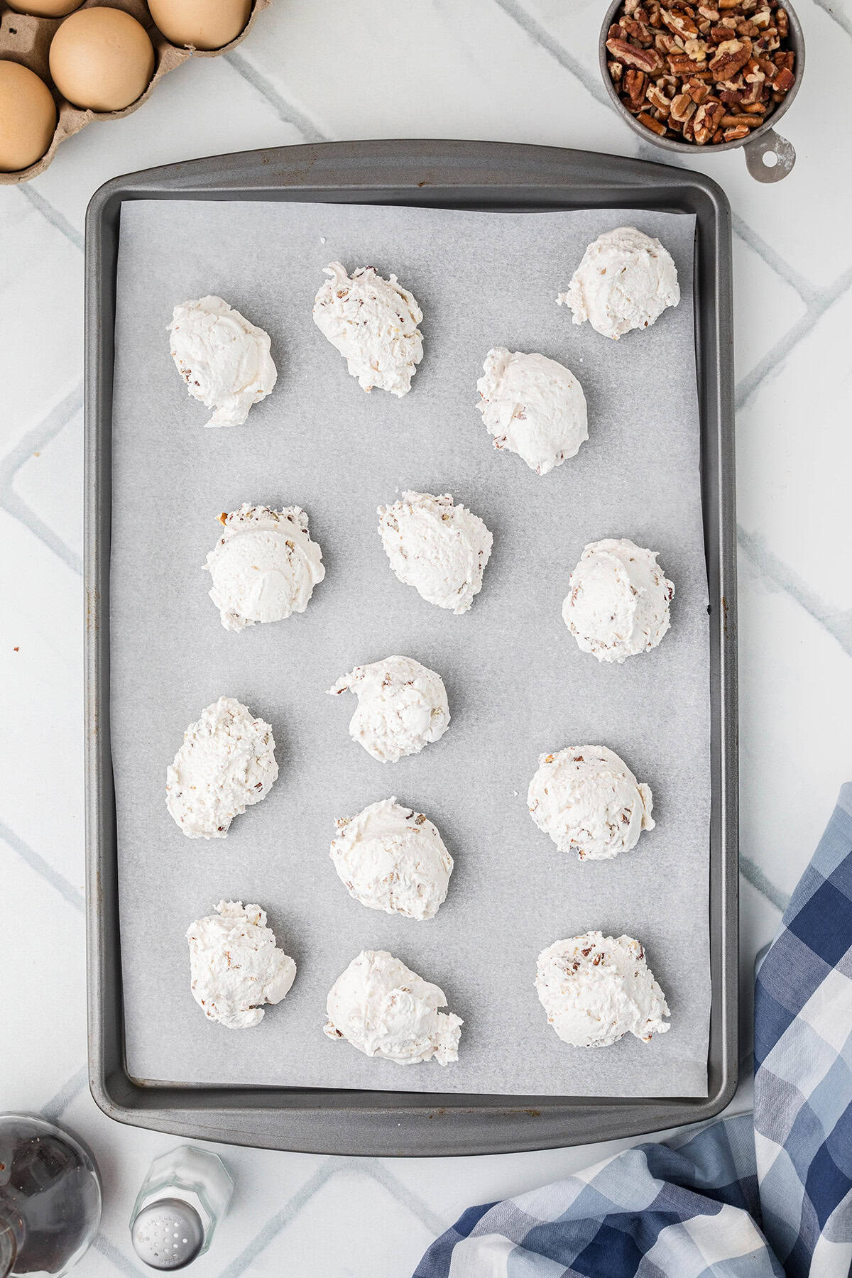 A baking sheet with 15 scoops of divinity candy on parchment paper, ready to bake. Eggs and pecans nearby.