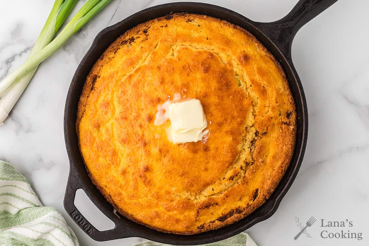 Golden cornbread in a cast iron skillet with melting butter on top, next to green onions on a marble surface.