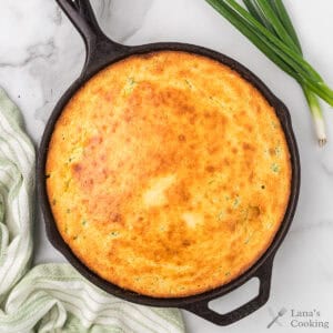 Golden cornbread baked in a cast iron skillet, with green onions and a striped towel beside it.