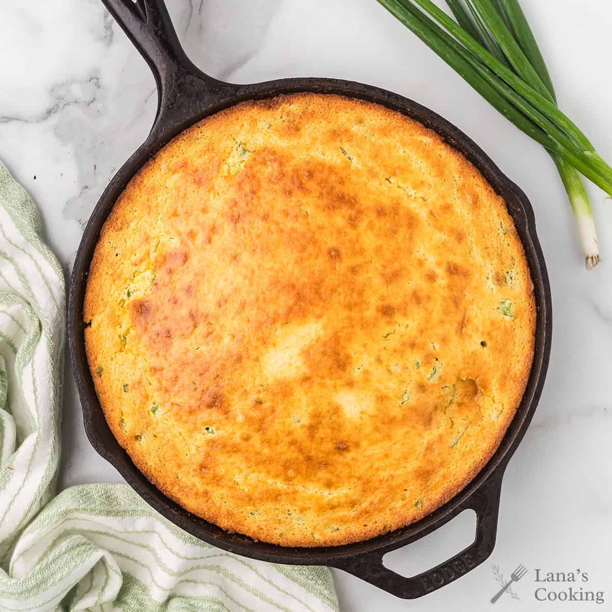 Golden cornbread baked in a cast iron skillet, with green onions and a striped towel beside it.