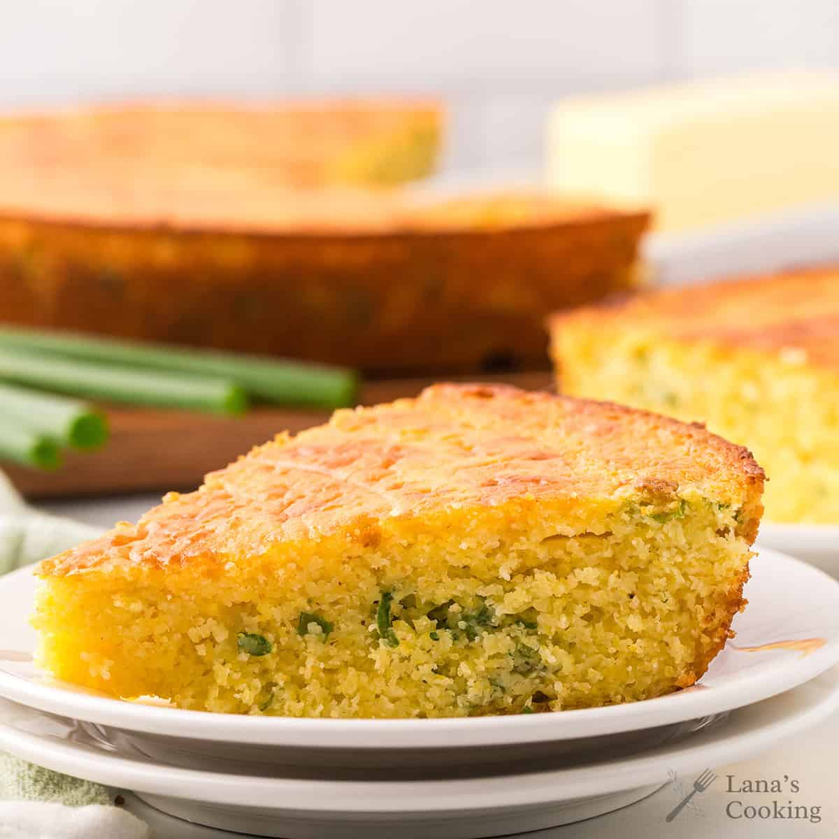 A slice of cornbread with green onions on a white plate, with more cornbread in the background.