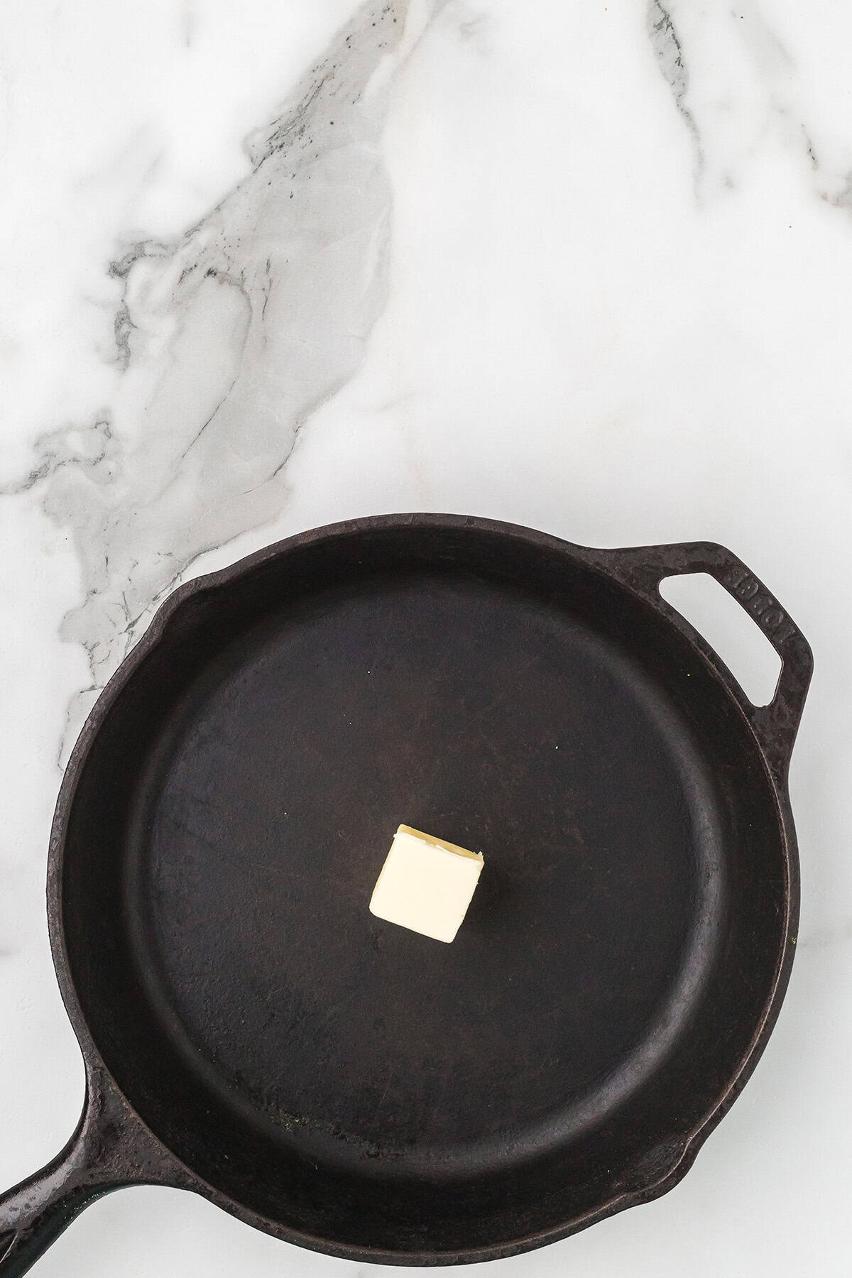 A small cube of butter sits in the center of an empty cast iron skillet on a marble surface.