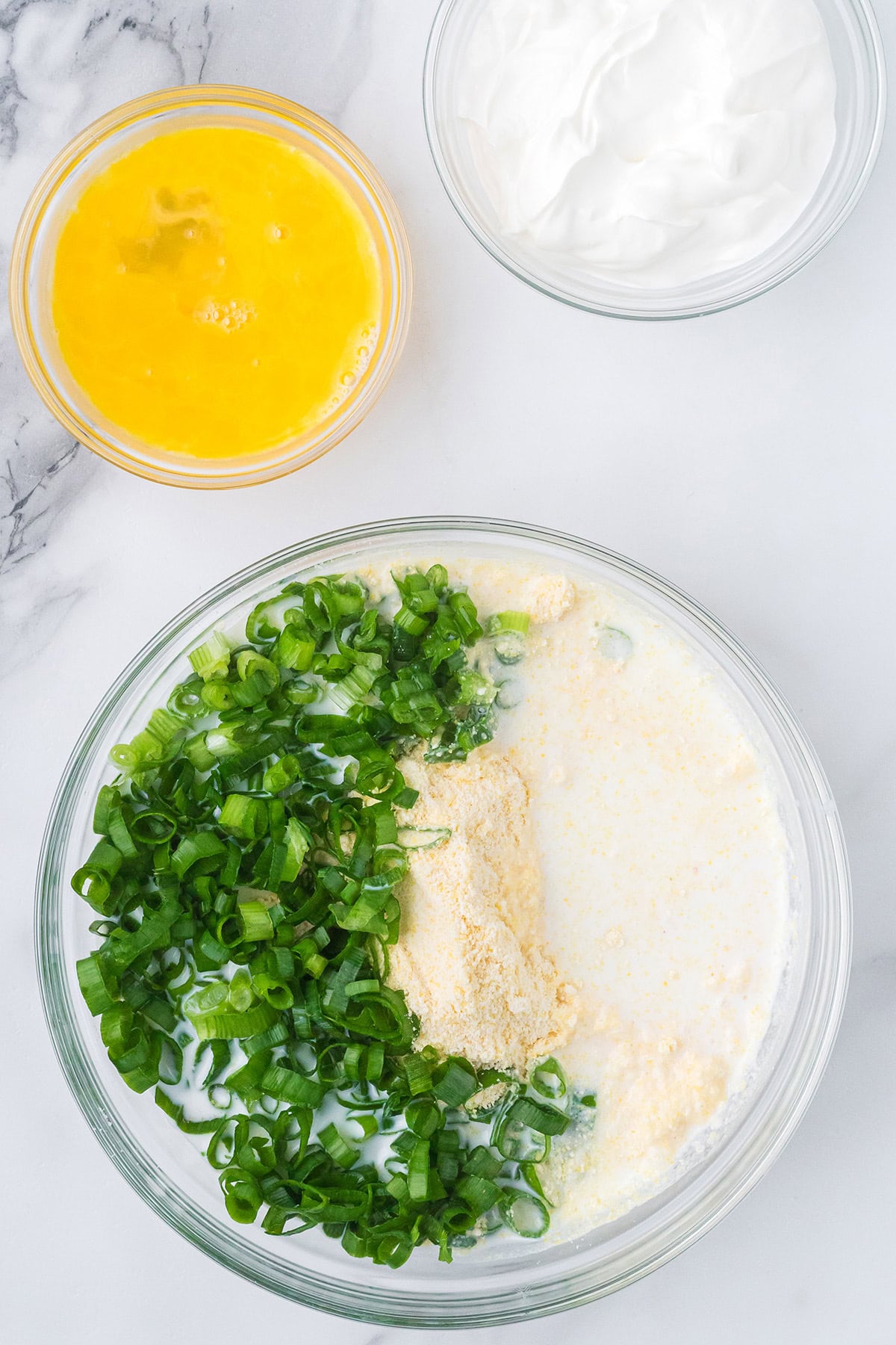 Three bowls: one with beaten eggs, one with sour cream, and one with chopped green onions, cornmeal, and milk.