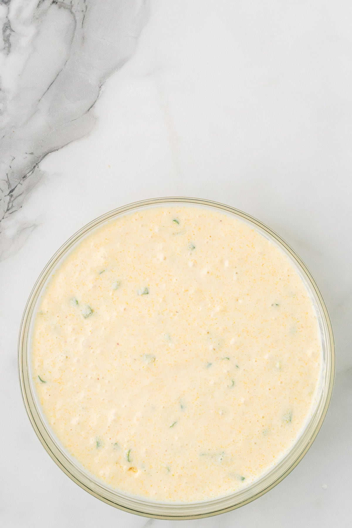 A glass bowl filled with creamy, yellow cornbread batter speckled with green herbs on a marble surface.