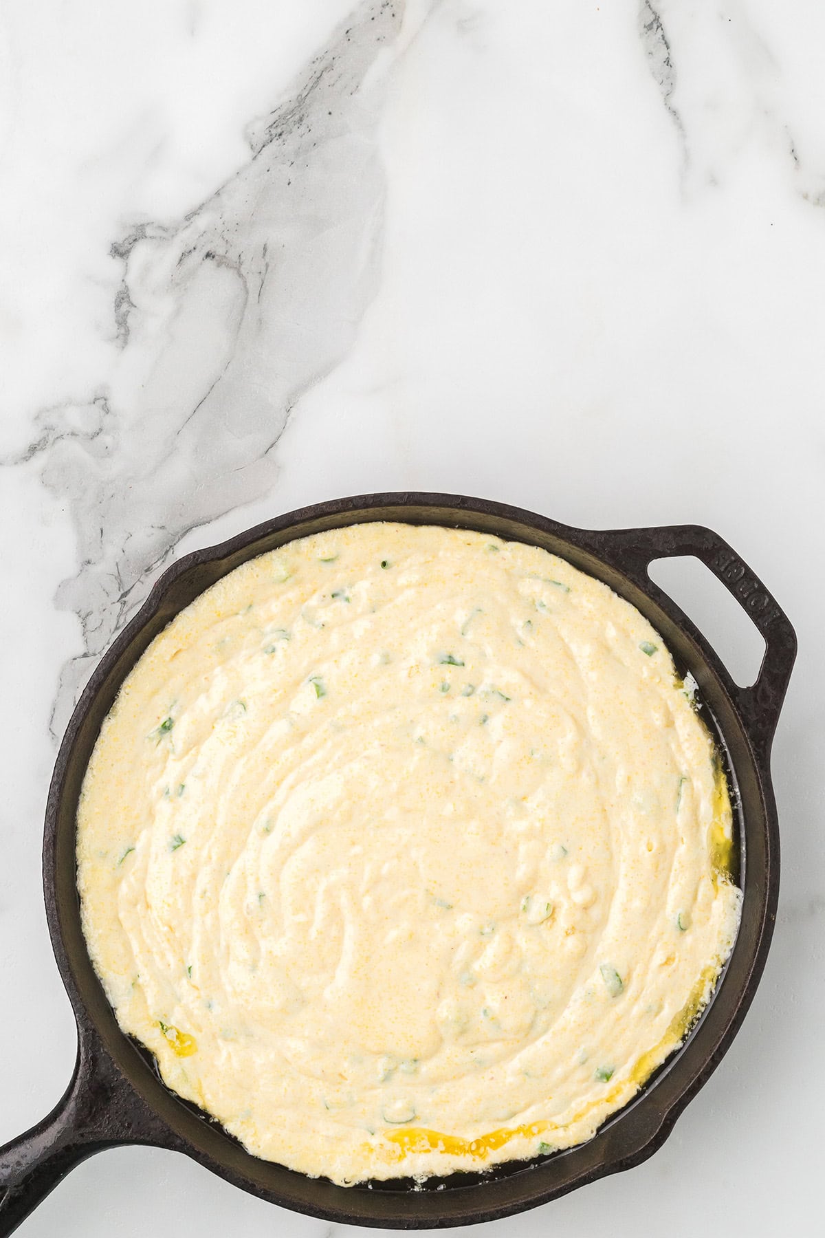 Cornbread batter with green herbs in a cast iron skillet on a white marble countertop.
