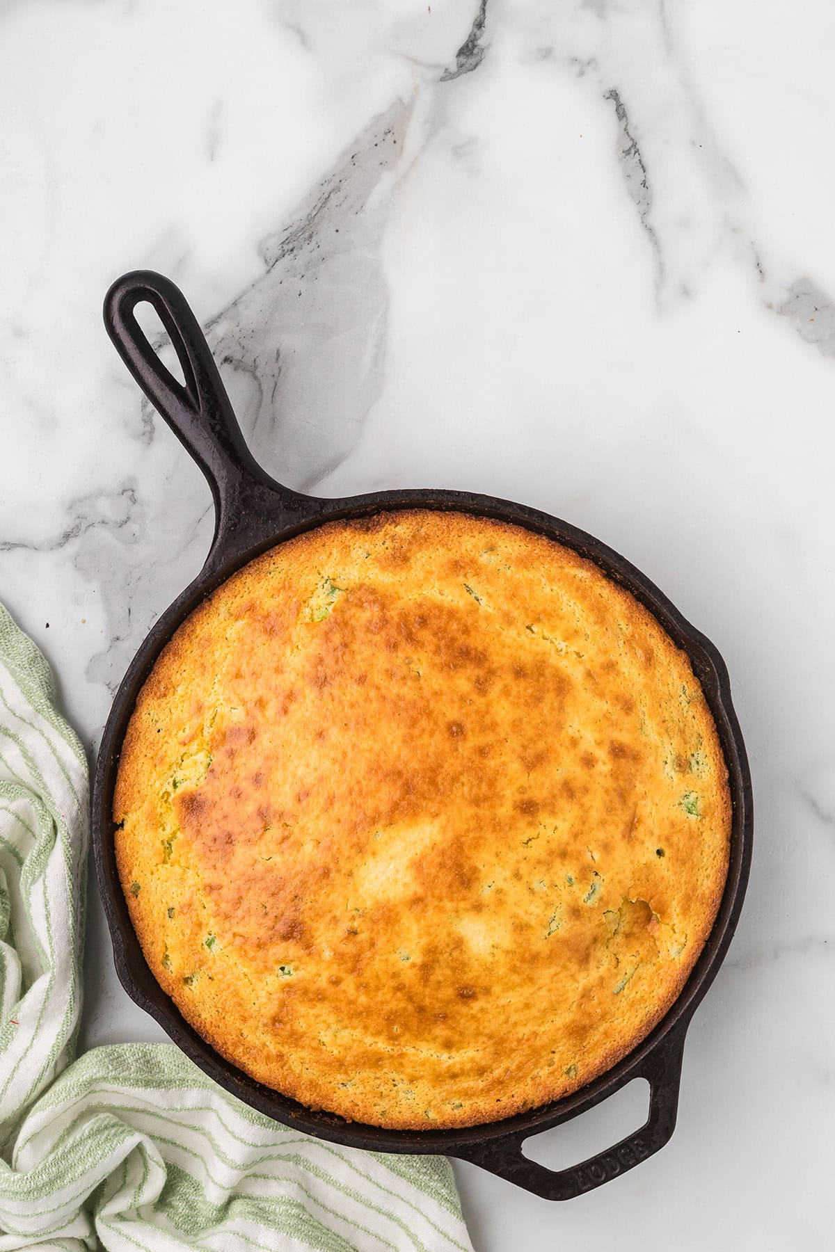 Golden cornbread baked in a black cast iron skillet on a marble surface, with a striped towel nearby.
