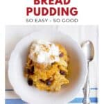 A bowl of bread pudding topped with whipped cream and raisins, next to a spoon on a striped cloth.