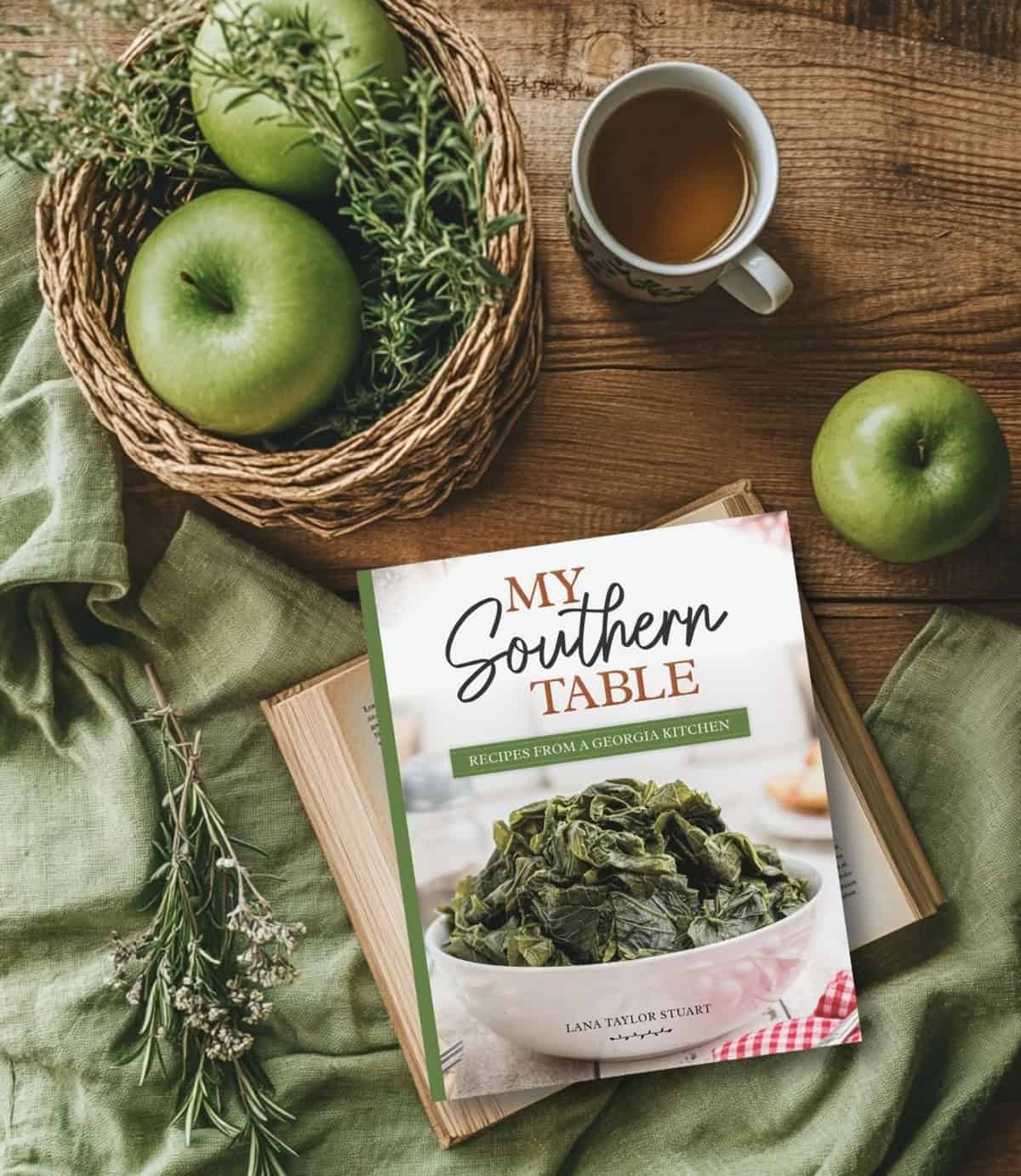 A cookbook, green apples in a basket, herbs, and a cup of tea arranged on a rustic wooden table.
