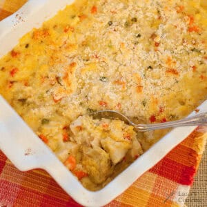 A spoon scoops creamy casserole with vegetables from a white baking dish on a colorful placemat.