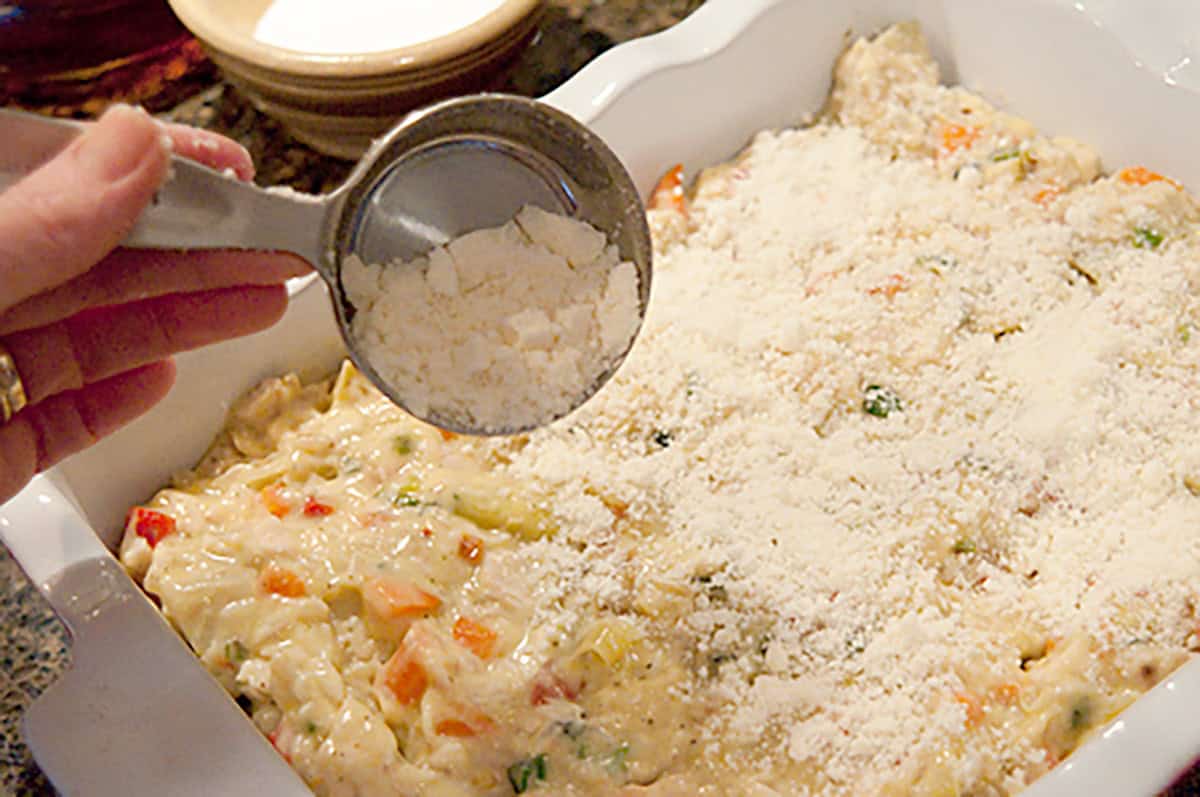 A hand sprinkles grated cheese over a casserole dish filled with a creamy vegetable mixture.