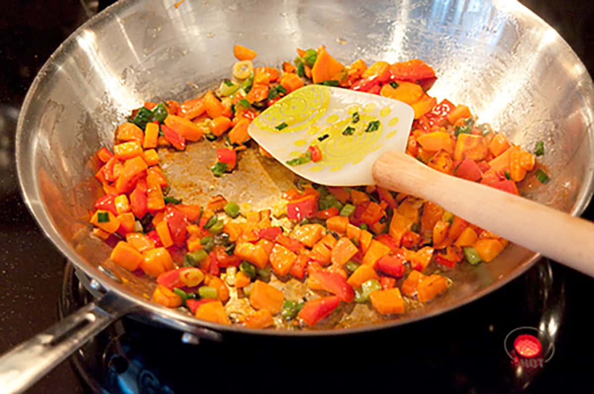 Diced vegetables saut&eacute;ing in a stainless steel pan with a spatula on a stovetop.