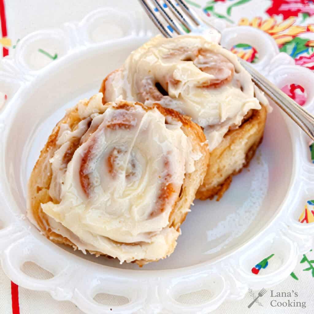 Two cinnamon rolls with creamy icing on a white plate, with a fork and floral napkin beside them.
