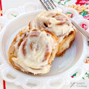 Two cinnamon rolls with creamy icing on a white plate, with a fork and floral napkin beside them.