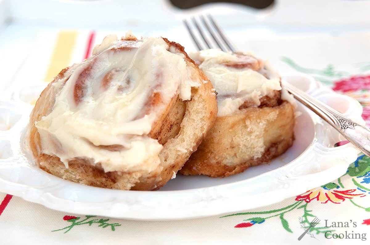 Two frosted cinnamon rolls on a white plate with a fork, set on a floral-patterned tablecloth.