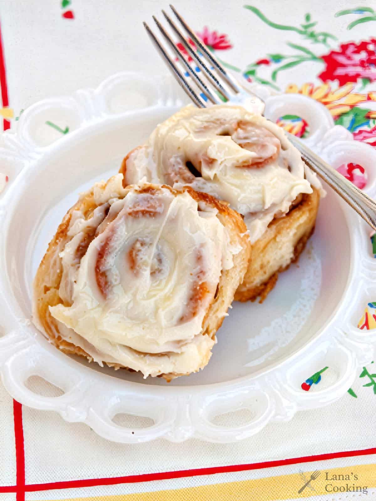 Two cinnamon rolls with white icing on a white plate, with a fork, on a colorful floral tablecloth.