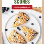 Three triangular lemon blueberry scones with icing, on a white plate, surrounded by blueberries and lemon.