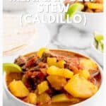 A bowl of Mexican beef stew with potatoes, tomatoes, lime, and tortillas on the side.