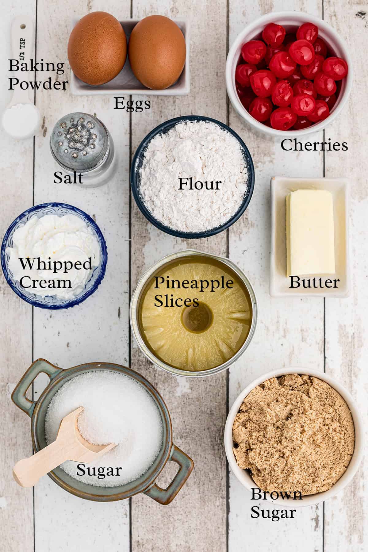 Ingredients on a table: eggs, cherries, flour, butter, salt, whipped cream, pineapple, sugar, brown sugar, baking powder.