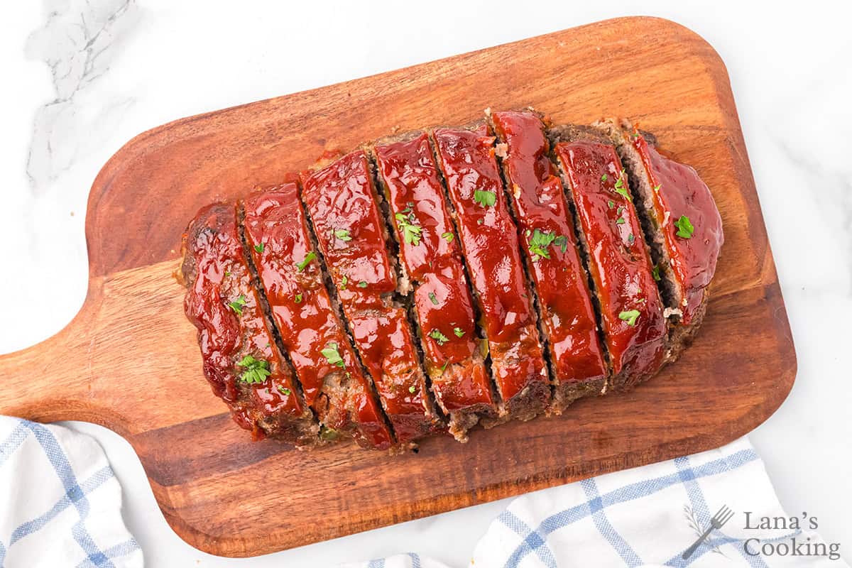 Sliced glazed meatloaf topped with herbs on a wooden cutting board.