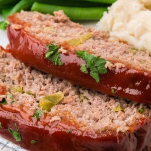 Two slices of glazed meatloaf garnished with parsley, served with mashed potatoes and green beans.
