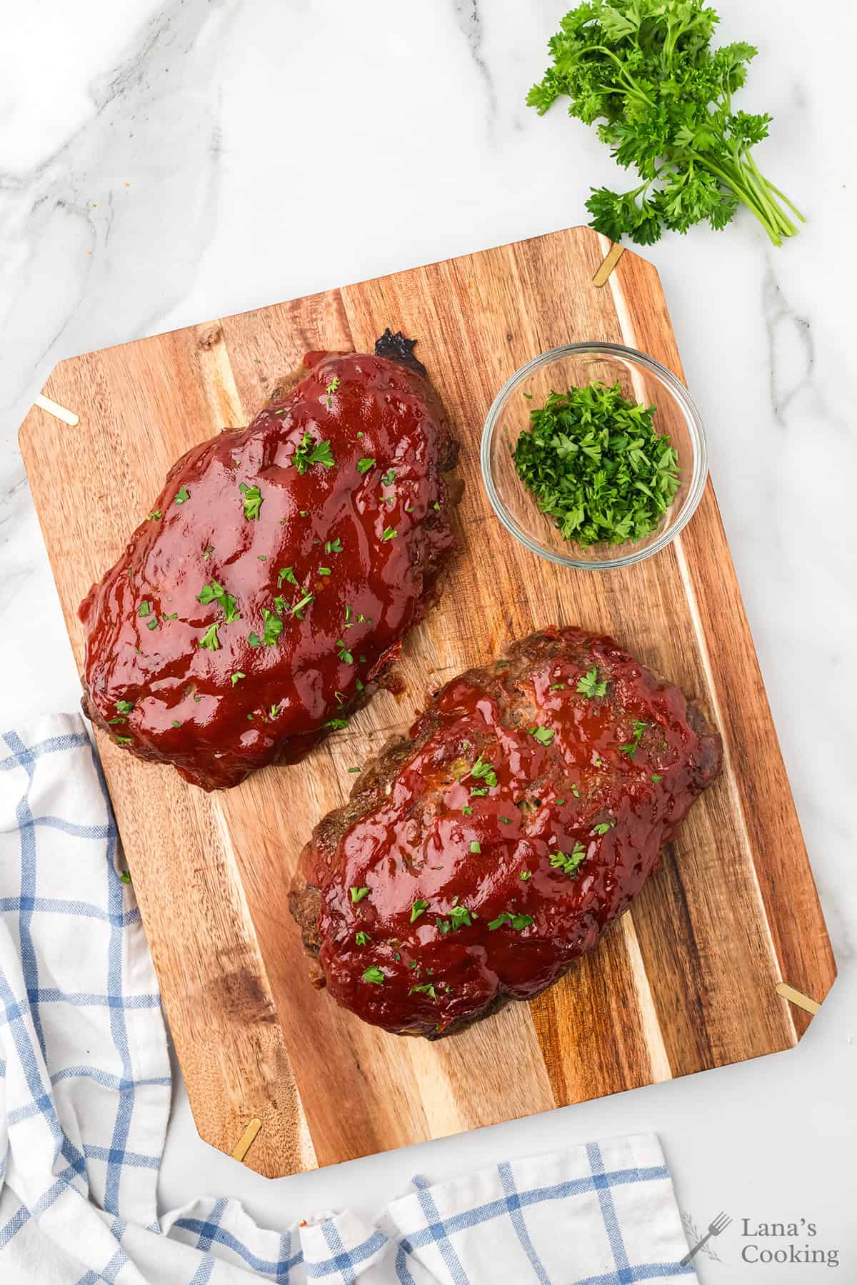 Two glazed meatloaves with parsley garnish on a wooden board, next to a small bowl of chopped parsley.
