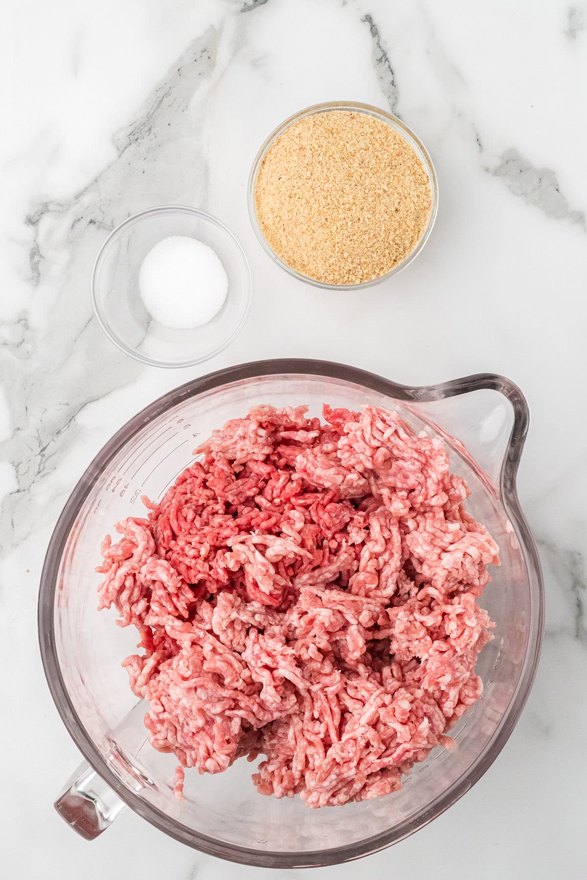 Ground meats in a glass bowl, with small bowls of breadcrumbs and salt on a marble countertop.