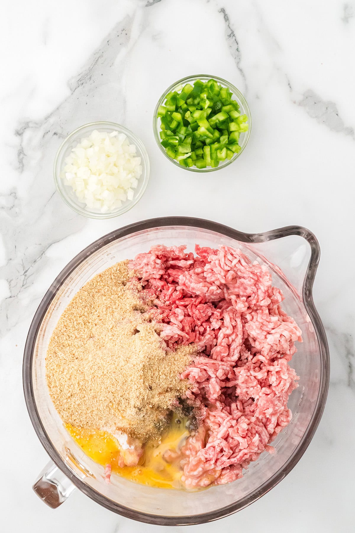 A bowl with ground meat, breadcrumbs, eggs, and seasonings, with chopped onion and green pepper in small bowls.