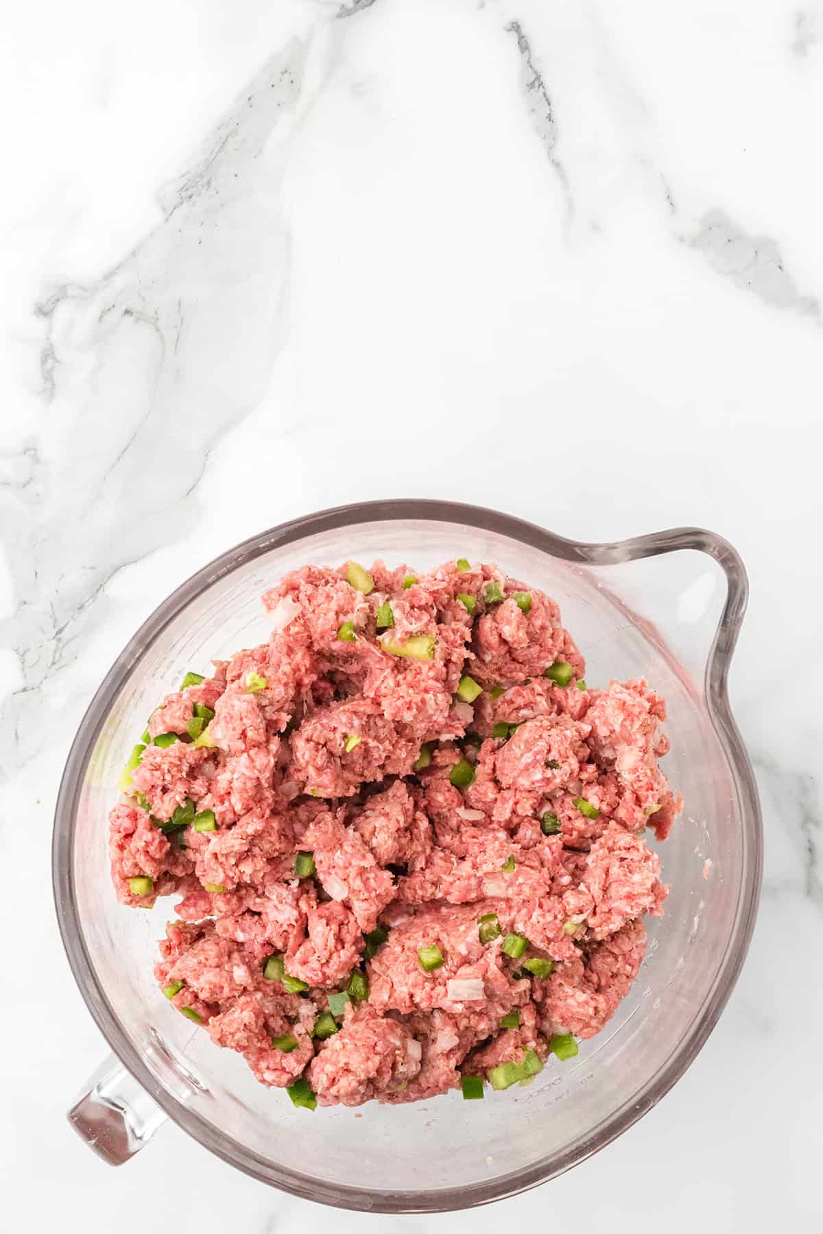 Ground meat mixed with chopped green peppers in a clear glass bowl on a white marble surface.