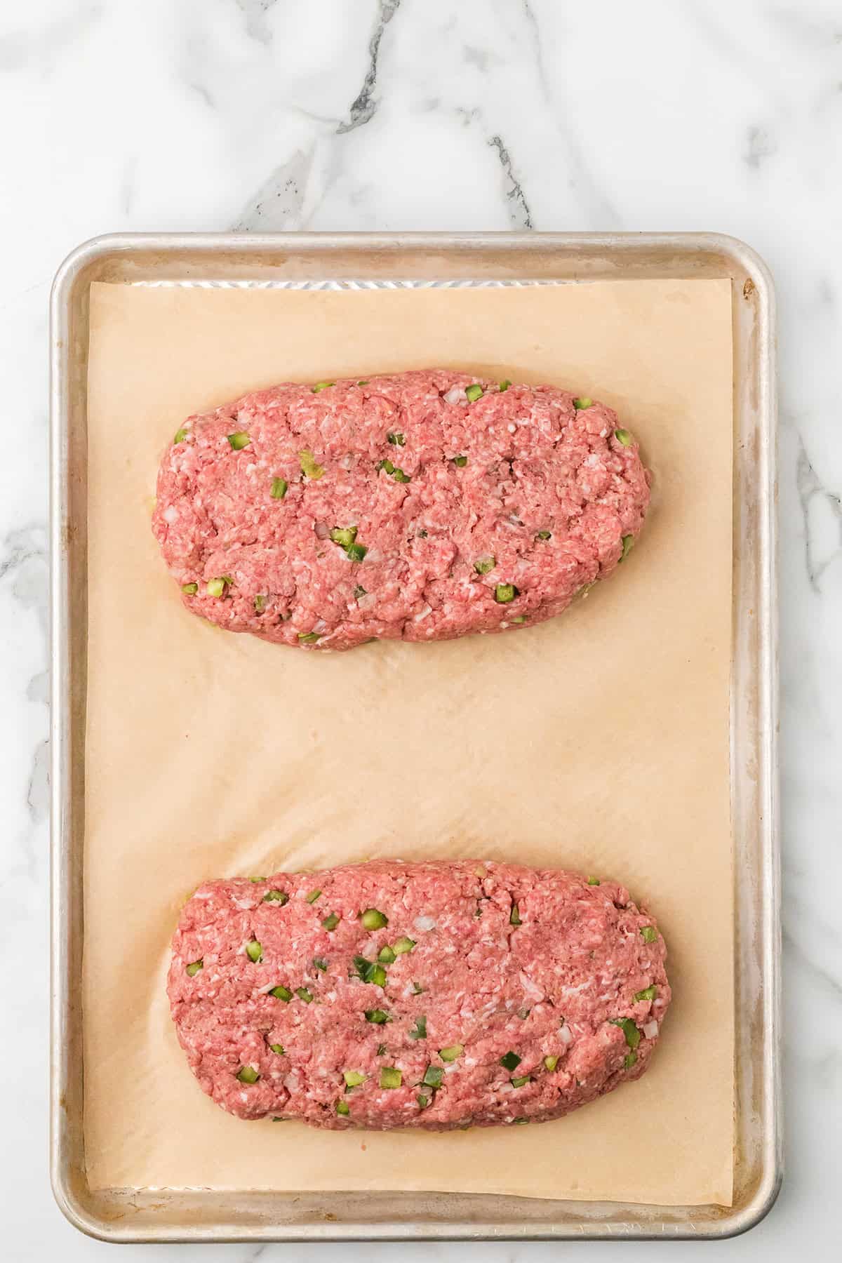 Two uncooked meatloaves with green bits on parchment paper on a baking sheet, on a marble surface.