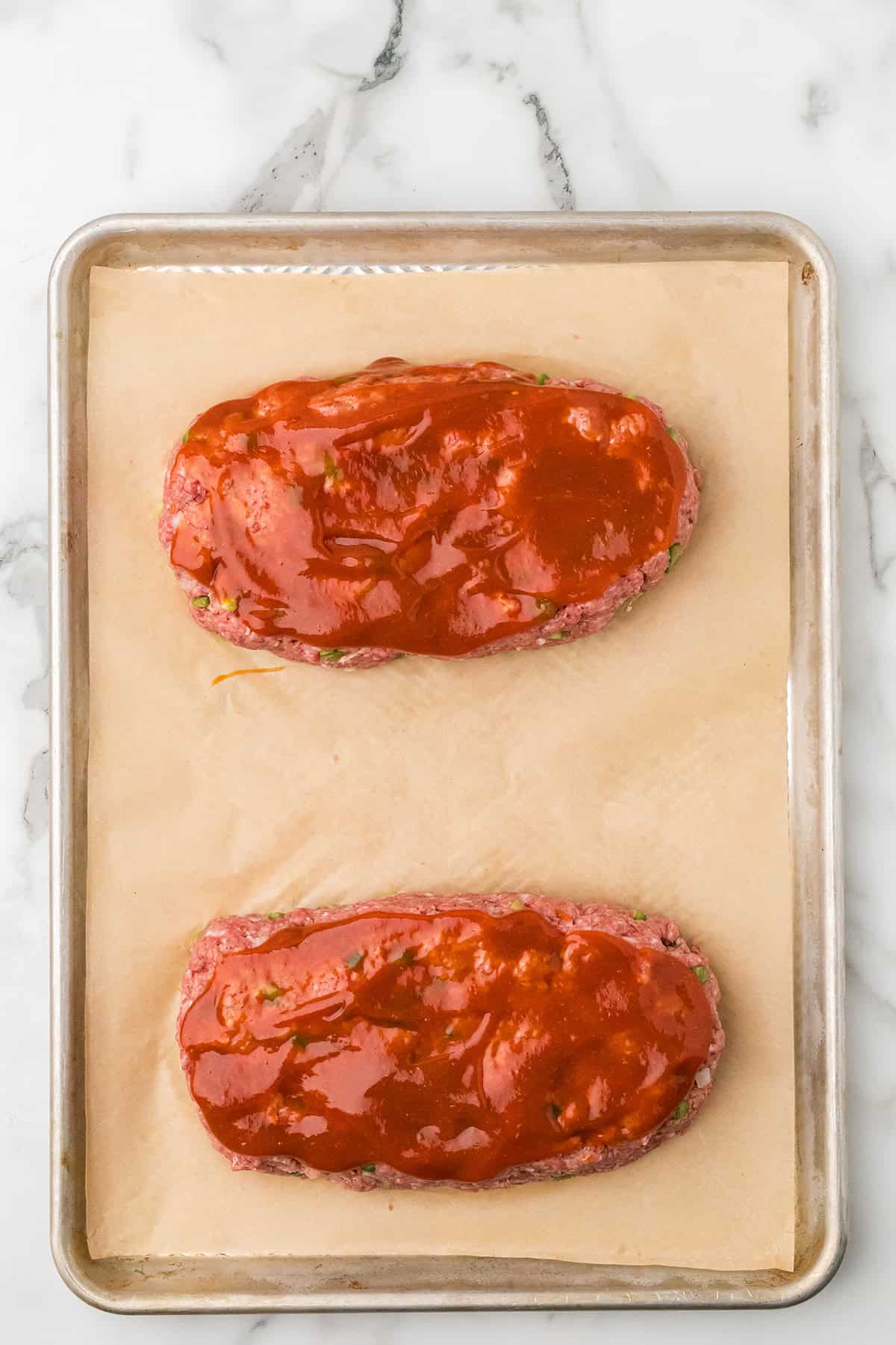 Two uncooked meatloaves topped with sauce on parchment paper on a baking sheet, viewed from above.