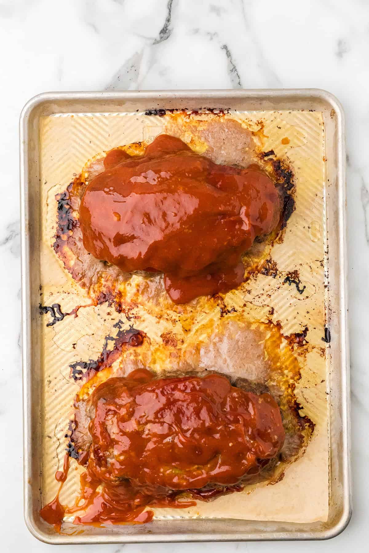 Two meatloaves topped with red sauce on a metal baking sheet, shot from above on a marble surface.